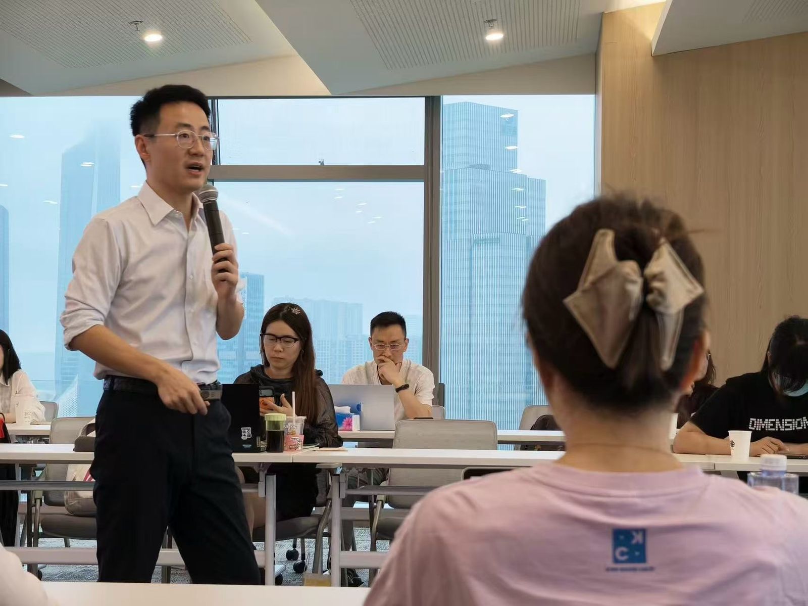工作坊现场,讲师持话筒面向参会者讲解,参会者在座位上使用电脑记录