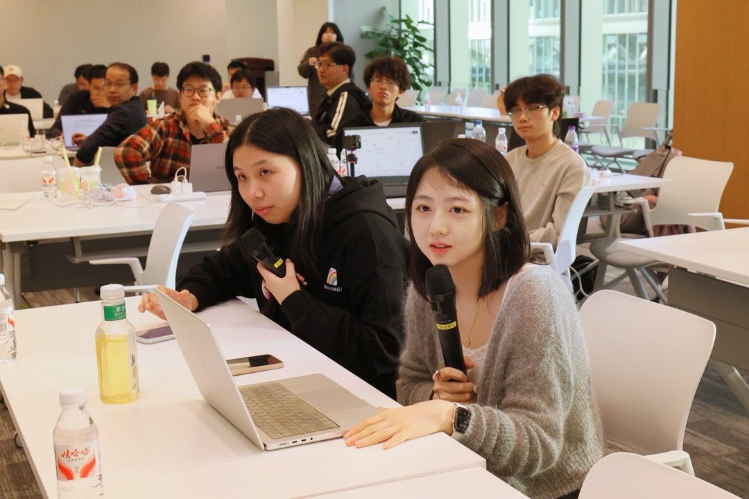 两位参赛者在桌前对着笔记本讲解作品,后排是旁听的参与者